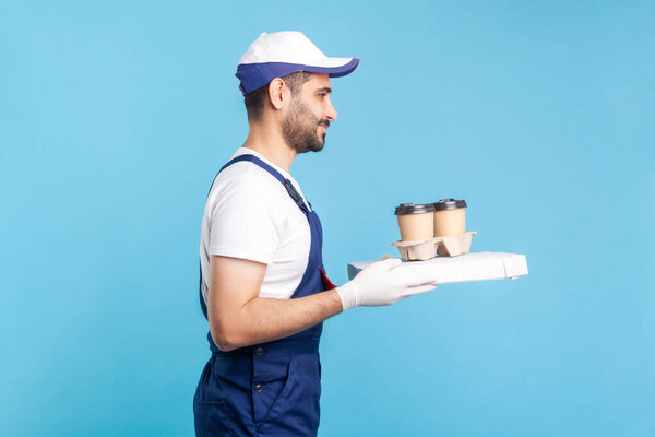 Delivery service. Side view, friendly professional courier in uniform holding coffee and pizza box, wearing safety gloves offering drinks and fast food, express shipping. indoor studio shot, isolated