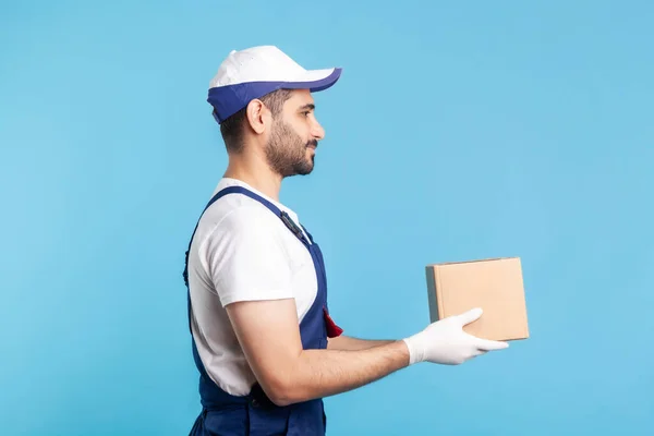 Delivery and cargo transportation. Side view, professional loader man in workwear holding cardboard box. Courier carrying parcel. Online express shipping, relocation moving services. studio shot