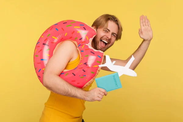Optimistic tourist guy in undershirt standing with pink donut rubber ring, holding passport document and airplane mockup, waving hello hi or goodbye gesture, hurry to travel. studio shot isolated