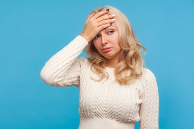 Üzgün, umutsuz, kıvırcık sarı saçlı bir kadın facepalm hareketi yapıyor, utanç duyuyor, başını belaya sokuyor, hata yapıyor. Mavi arka planda kapalı stüdyo çekimi