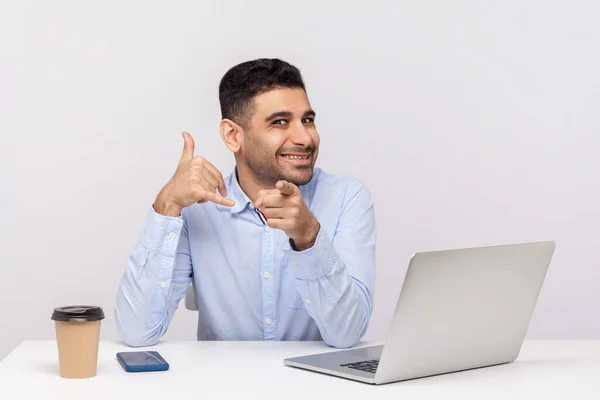 Hey you, let\'s contact by phone! Happy man office worker sitting at workplace, pointing to camera, keeping hand shaped like telephone, doing call me gesture. studio shot isolated on white background