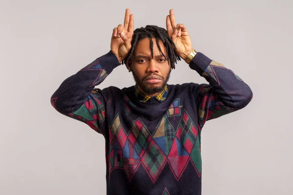 Upset frustrated african guy holding arms above head showing horns gesture, angry disrespectful offender seriously looking at camera. Indoor studio shot isolated on gray background