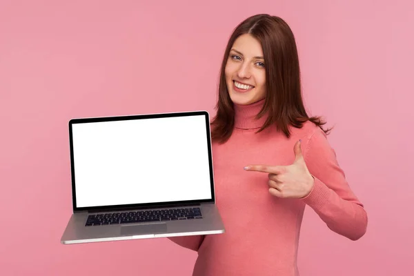 彼女が手に持っている空白のディスプレイでノートパソコンで指を指しているブルネットの女性は 空の画面を示し インターネット広告で自慢しています ピンクの背景に隔離された屋内スタジオショット — ストック写真