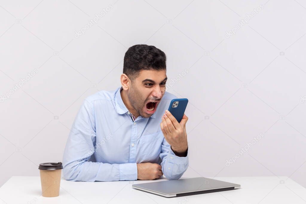 Mad crazy man employee sitting in office workplace, shouting furious aggressive at smartphone, pissed off after mobile talk, consulting annoying client. indoor studio shot isolated on white background