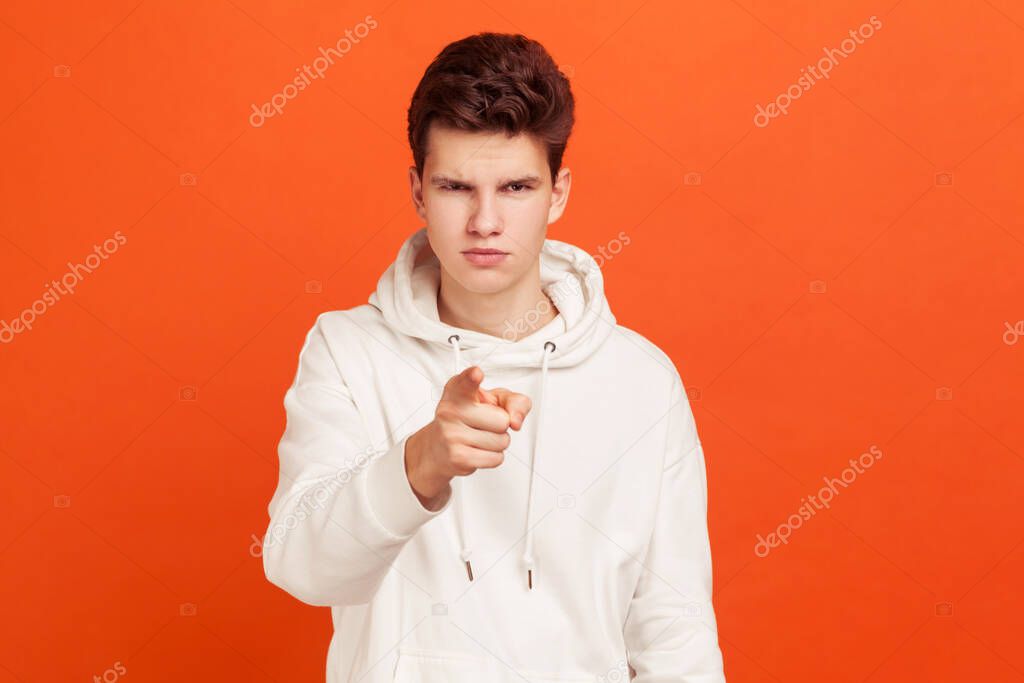 Confident young man with stylish hairdo in casual hoodie pointing finger on you and looking atcamera. You are next. Indoor studio shot isolated on orange background