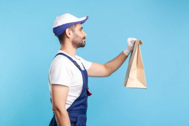 Yan görüş, üniformalı dost canlısı profesyonel kurye ve güvenlik eldivenleri, alışveriş torbası, paket yemek teslimatı, posta servisi ekspres nakliyesi. Mavi arka planda kapalı stüdyo çekimleri