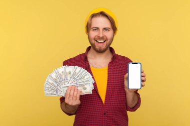 Çevrimiçi bankacılık, cep telefonu para transferi. Damalı tişörtlü neşeli hippi, elinde dolar banknotları ve boş ekranlı cep telefonu ile reklam için makara yapıyor. Sarı renkte kapalı stüdyo çekimi.