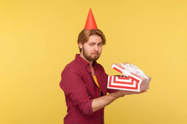 Kareli gömlekli mutsuz üzgün adam ve parti konisi hediye kutusu, kameraya bakıyor hayal kırıklığına uğramış, hatalı doğum günü hediyesi yüzünden üzgün. Sarı arka planda izole edilmiş kapalı stüdyo çekimi