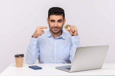 Kripto para, dijital para. Gömlekli şık bir adam iş yerinde dizüstü bilgisayarla oturuyor, altın bitcoin doğrultuyor, elinde btc sikke gösteriyor. Beyaz arka planda izole edilmiş kapalı stüdyo çekimi