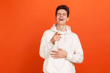 Beyaz kapüşonlu mutlu genç adam seni işaret ediyor ve içten bir şekilde el ele gülüyor. Eğleniyorum. Turuncu arka planda kapalı stüdyo çekimleri