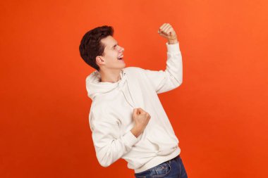 İşte bu! Şık, rahat kapüşonlu genç son derece memnun. Zaferini kutluyor ve evet hareketi yapıyor, olumlu duygular gösteriyor. Turuncu arka planda kapalı stüdyo çekimleri