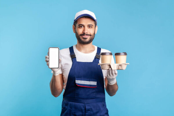 Delivery mobile app, internet advertise. Happy friendly courier man in overalls holding coffee cups and cellphone with mock up display, online order service. studio shot isolated on blue background