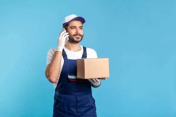 Delivery service. Courier answering phone call, holding cardboard box and talking with client, carrying online order in parcel. Express shipping, relocation service. indoor studio shot isolated