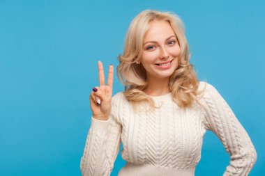 Tatmin olmuş sarışın kadın kameraya bakarken dişlek gülümseme, huzur, zafer sembolü, kazanmasıyla ve kutlamalarıyla gurur duyuyor. Mavi arka planda kapalı stüdyo çekimi