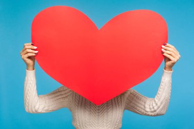 Kırmızı kağıt kalbin, hayır kurumunun, yardım ve desteğin, sağlık hizmetleri ve organ nakli organizasyonunun arkasına saklanan beyaz örülü kazaklı bir kadın. Mavi arka planda kapalı stüdyo çekimi