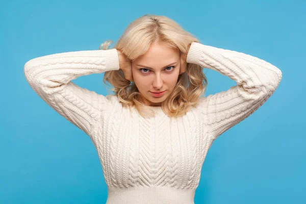 Irritated nervous blond woman in white sweater closing ears with hands, lady in despair avoiding noisy sound, headache. Indoor studio shot isolated on blue background