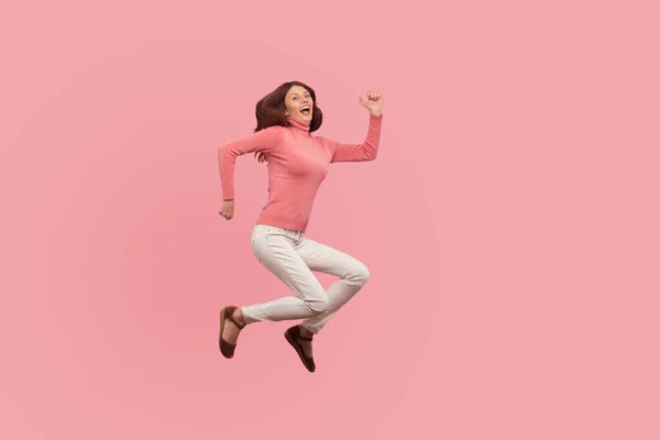 非常に幸せな茶色の髪の高いジャンプや空気中を飛んで満足し 女性は 夢が叶う ピンクの背景に隔離された屋内スタジオショット — ストック写真