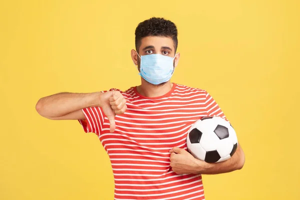 Upset man in medical safety mask and in red t-shirt holding ball showing dislike, cancellation of championship due to coronavirus epidemic. Indoor studio shot isolated on yellow background