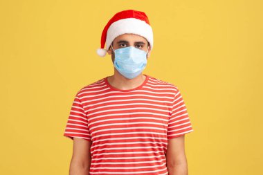 Kırmızı tişörtlü, Noel Baba şapkalı, yüzünde cerrahi maske olan ve kameraya bakan ciddi bir adam. Sarı arka planda kapalı stüdyo çekimleri