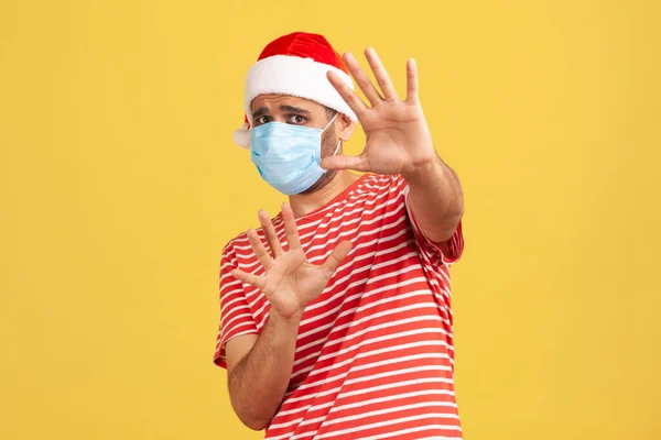 Scared man in medical protective face mask in red santa claus hat holding hands raised, afraid of covid-2019 virus during holidays, shock and panic. Indoor studio shot isolated on yellow background