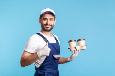 Teslimat servisi. Yan görüş, tulumlu arkadaş canlısı kurye elinde kahve, güvenlik eldiveni takıyor tek kullanımlık bardaklarda içki ikram ediyor ve baş parmağını kaldırıyor. Kapalı stüdyo çekimi, izole.