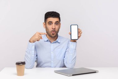 Ofis işyerinde oturan hayret verici işadamı, cep telefonuna boş ekranla bakıyor ve şaşkın şaşkın bakıyor, reklam için boş bir ekran tutuyor. Stüdyoda çekim izole edildi