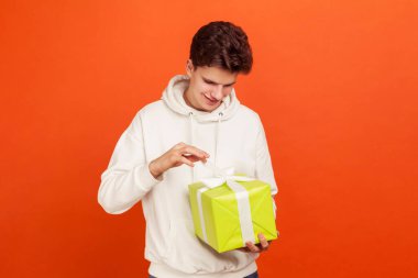 Handsome young man in casual white sweatshirt with hood adjusting bow on giftbox and smiling, teenager preparing for date, birthday gift. Indoor studio shot isolated on orange background clipart