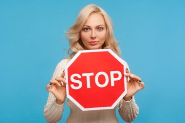 Serious concentrated woman with blond hair holding in hands stop sign, warning about danger, denies bad habits. Indoor studio shot isolated on blue background clipart