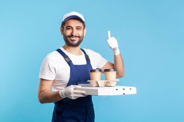 Delivery service. Friendly optimistic courier in overalls holding coffee and pizza box, wearing safety gloves offering drinks food and showing thumbs up, like gesture. indoor studio shot, isolated