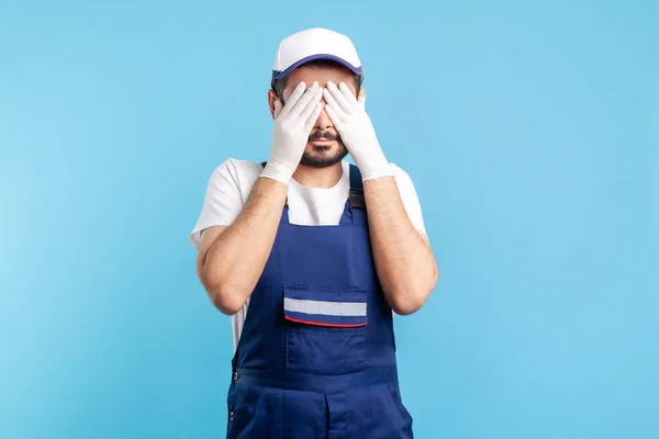 Don't want to look! Handyman in overalls and hygiene gloves covering eyes, refusing to watch. Profession of service industry, courier delivery, housekeeping maintenance. indoor studio shot isolated