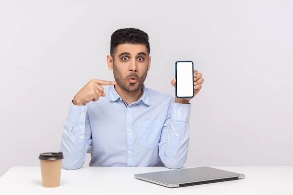 Amazed businessman sitting in office workplace, pointing at mobile phone with empty screen and looking surprised shocked, holding mock up blank display for advertisement. indoor studio shot isolated