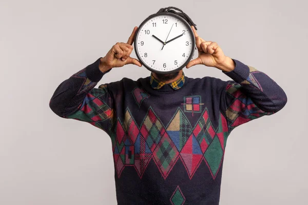 Time to work! African man hiding face behind big wall clock, afraid of deadline, wasting time, being late. Indoor studio shot isolated on gray background
