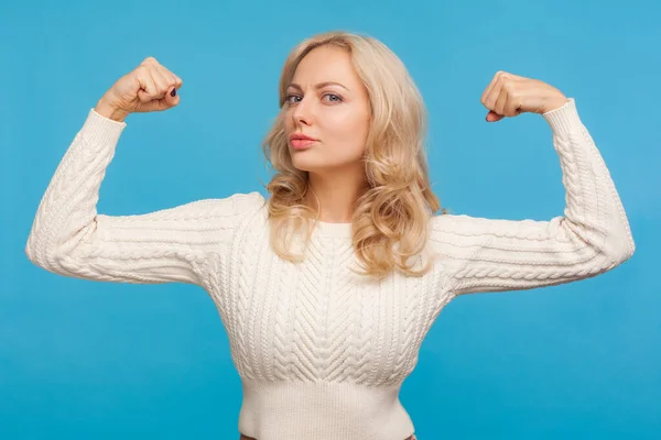 Strong independent woman with blond hair looking at camera showing her arm muscles, proud of her strength and leadership skills, emancipation. Indoor studio shot isolated on blue background