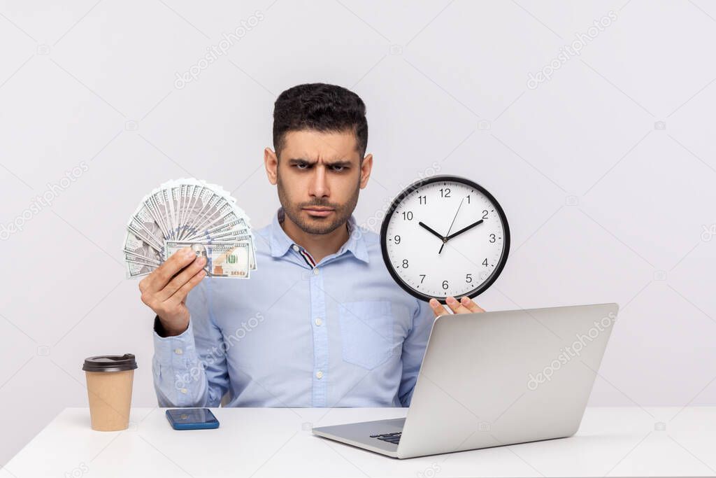 Time is money! Bossy angry businessman sitting in office workplace, holding big clock and dollar banknotes, frowning looking at camera strict expression. studio shot isolated on white background
