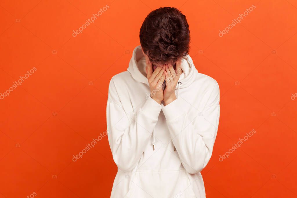 Upset lonely teenager in hoodie closing face with palms crying with despair, hiding his eyes in shame, depression. Indoor studio shot isolated on orange background