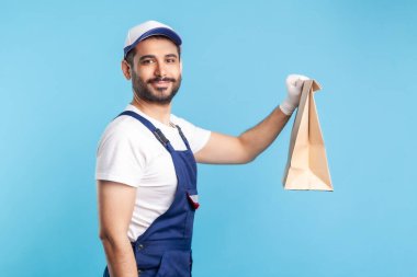 Yan görüş, üniformalı mutlu profesyonel kurye ve güvenlik eldivenleri yiyecek torbası veriyor, paket yemek dağıtıyor ve kameraya gülümsüyor, posta servisi ekspres nakliyesi yapıyor. Kapalı stüdyo çekimi, izole.