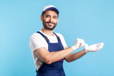 Tulum giymiş, ameliyat eldivenleriyle elini gösteren ve kameraya gülümseyen mutlu tamirci. Hizmet sektöründe güvenlik giysileri, koruma ve hijyen, kurye teslimatı, ev bakımı. Stüdyo çekimi