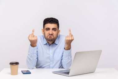Öfkeli kaba işadamı masa başında dizüstü bilgisayarla işyerinde oturuyor, orta parmağını gösteriyor, agresif bir şekilde bakıyor, saygısızlık ve nefret gösteriyor. Beyaz arka planda izole edilmiş kapalı stüdyo çekimi
