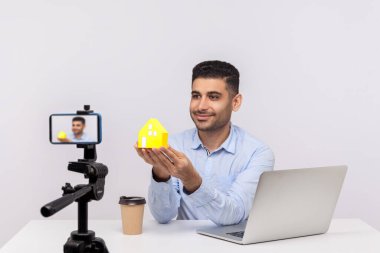 Pozitif gülümseyen adam, emlakçı ofis işyerinde oturuyor ve kağıt evi kameraya gösteriyor, video kaydediyor, satın alma ya da kiralama konutları hakkında çevrimiçi blog yayınlıyor. Stüdyo çekimi izole