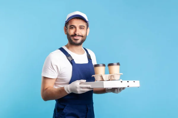 Delivery service. Friendly professional courier in uniform holding coffee and pizza box, wearing safety gloves offering drinks food and looking at camera with toothy smile. studio shot isolated