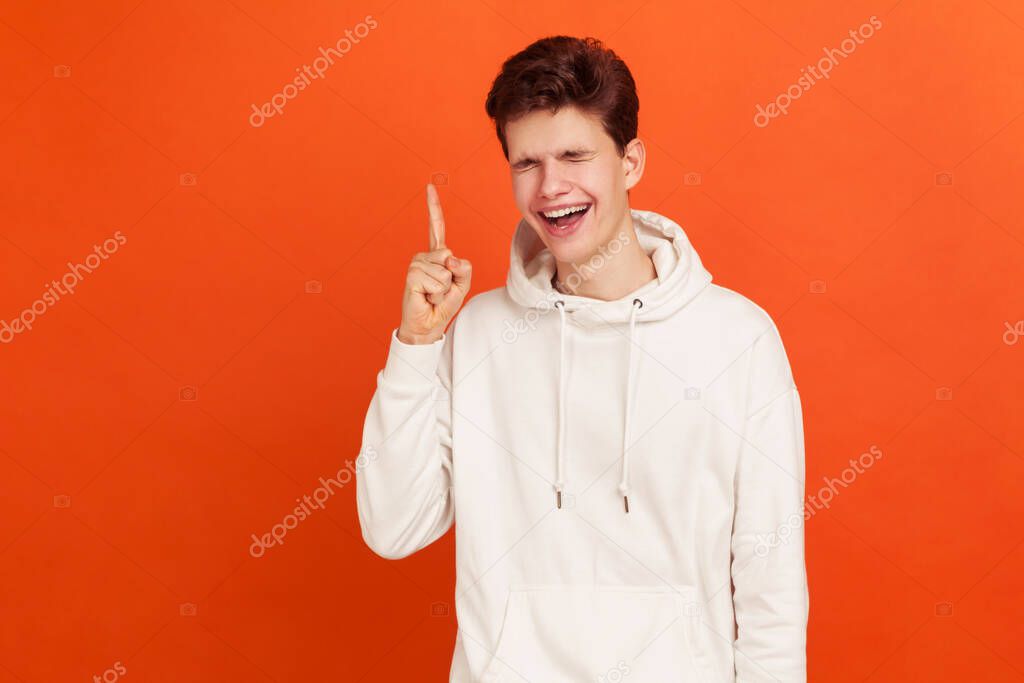 Cheerful young man pointing finger up and sincerely laughing, miscievous teenager thinking devious tricks and cheats. Indoor studio shot isolated on orange background