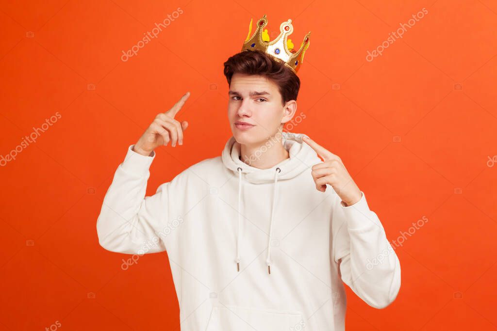 Selfish egoistic teenager in casual style white hoodie pointing fingers on golden crown on his head, showing his authority. Indoor studio shot isolated on orange background