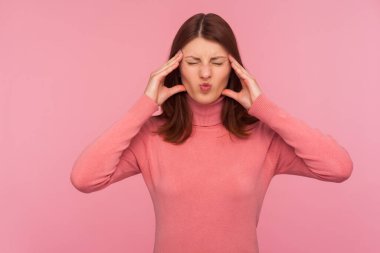 Pembe süveter giyen üzgün esmer kadın elleriyle şakak masajı yapıyor. Baş ağrısı çekiyor, migren riski var. Pembe arka planda kapalı stüdyo çekimleri