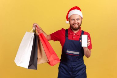 Noel Baba şapkalı ve mavi üniformalı mutlu kurye alışveriş çantaları ve akıllı telefon taşıyor dişlek gülümsemeli kameraya bakıyor, online mağaza. Sarı arka planda kapalı stüdyo çekimleri