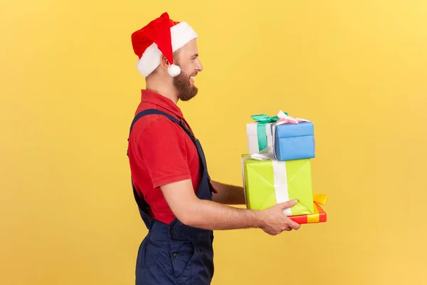Profile portrait male courier in professional uniform and red santa claus hat holding and giving presents, delivery in holidays. Indoor studio shot isolated on yellow background