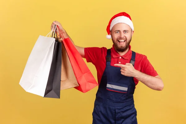 サンタクラスの帽子の陽気な正配達の男と彼の手のショッピングバッグで統一された指差し指 店からの迅速な配達 黄色の背景に隔離された屋内スタジオショット — ストック写真