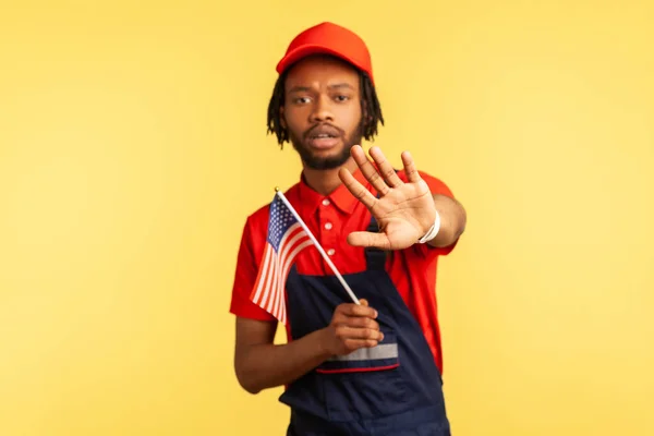 Serious afro-american craftsman with dreadlocks in uniform holding us flag showing stop gesture raising arm, no to racism, protection of human rights. Indoor studio shot isolated on yellow background
