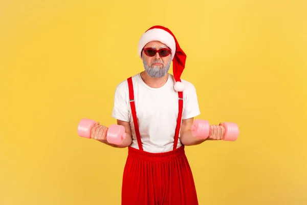Self confident elderly man in santa claus hat and sunglasses holding dumbbells, pumping muscles before holidays, having fun. Indoor studio shot isolated on yellow background