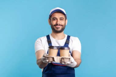 Teslimat servisi. Mavi tulumlu arkadaş canlısı kurye kahve ikram ediyor, güvenlik eldiveni giyiyor tek kullanımlık bardaklarda içki veriyor ve gülümseyerek kameraya bakıyor. Kapalı stüdyo çekimi, izole.