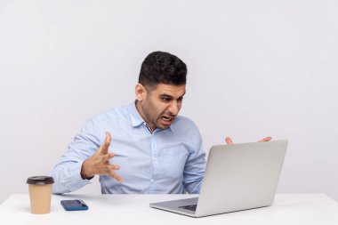 Sinirli işadamı ofis işyerinde oturmuş, öfkeli bir ifadeyle dizüstü bilgisayara bakıyor, çevrimiçi sohbet ederken bağırıyor, video görüşmesi yapıyor. Stüdyo çekimi beyaz arkaplanda izole edildi